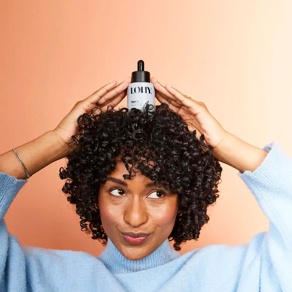Curly girl posing with a bottle of LOHY Hush Nourishing Oil on her head